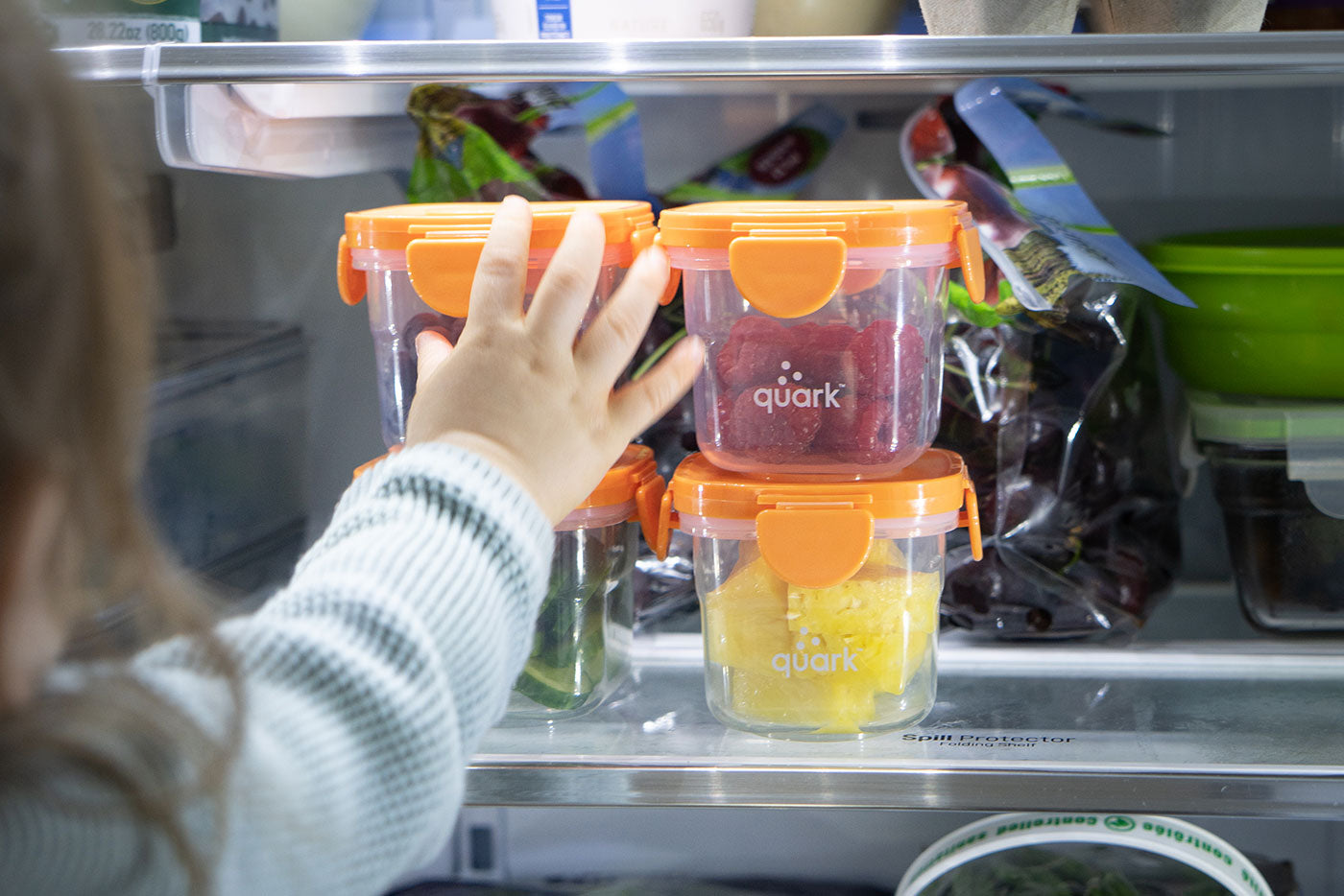 Child's hand reaching for Quark Baby orange storage containers with colorful food purees in refrigerator.