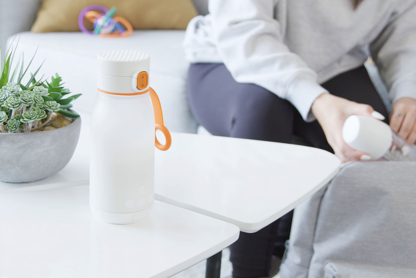 Quark Baby BuubiBottle portable milk warmer with orange handle displayed on white desk with succulent plant.