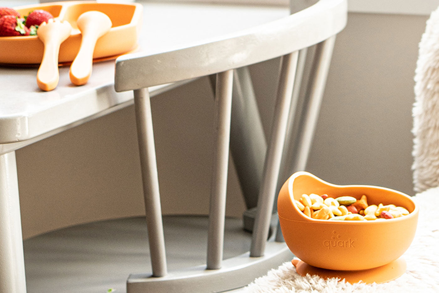 Quark Baby orange bowls with handles filled with colorful baby snacks and cereal on white pedestal table.