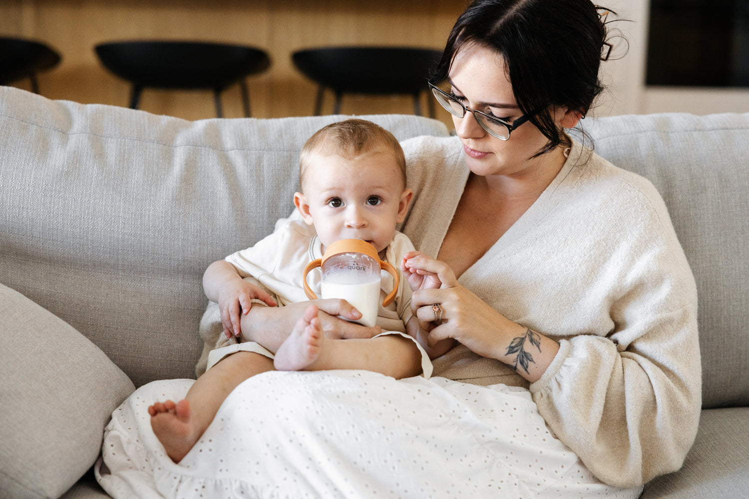 Mother holding Quark Baby sippy cup while feeding infant on couch during breastfeeding moment