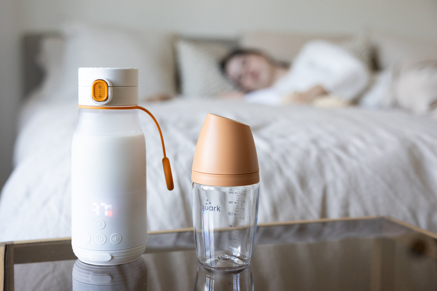 Quark Baby bottle and feeding cup displayed on nightstand beside sleeping infant in bedroom.