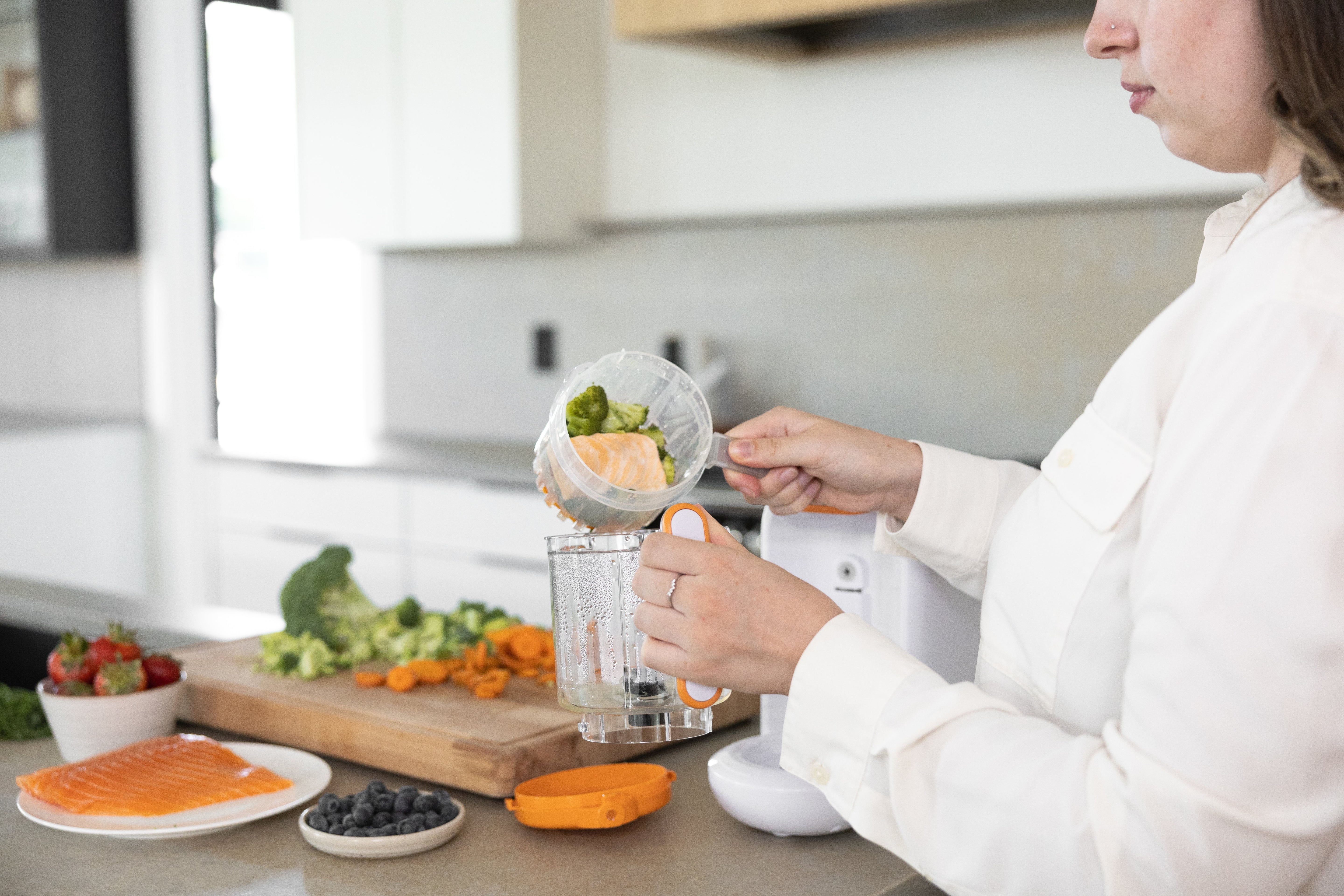 Woman blending fresh vegetables and broccoli with Quark Baby Quook food maker in modern kitchen.