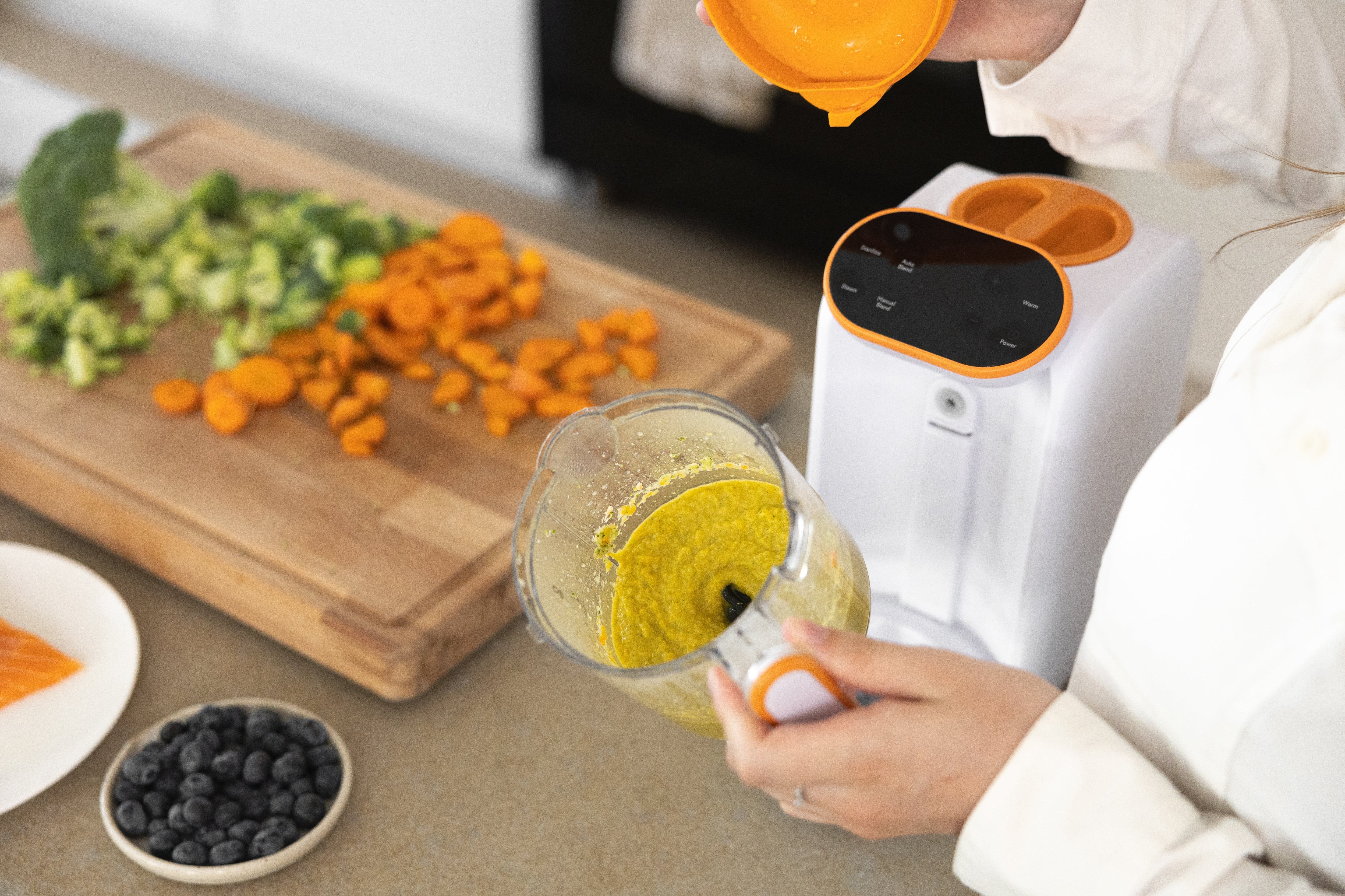 Adult hand squeezing fresh lemon into Quark Baby blender with chopped vegetables and blueberries nearby.