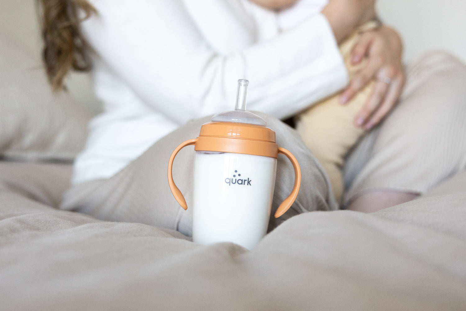 Adult holding Quark Baby orange sippy cup with handles while feeding infant in newborn routine lifestyle scene.