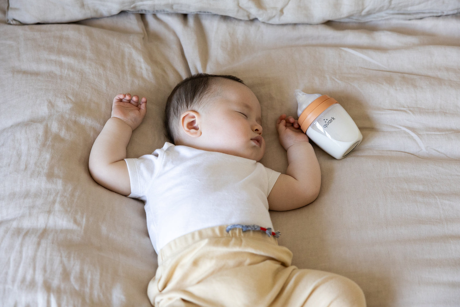 Sleeping newborn in white shirt next to Quark Baby bottle on beige bedding