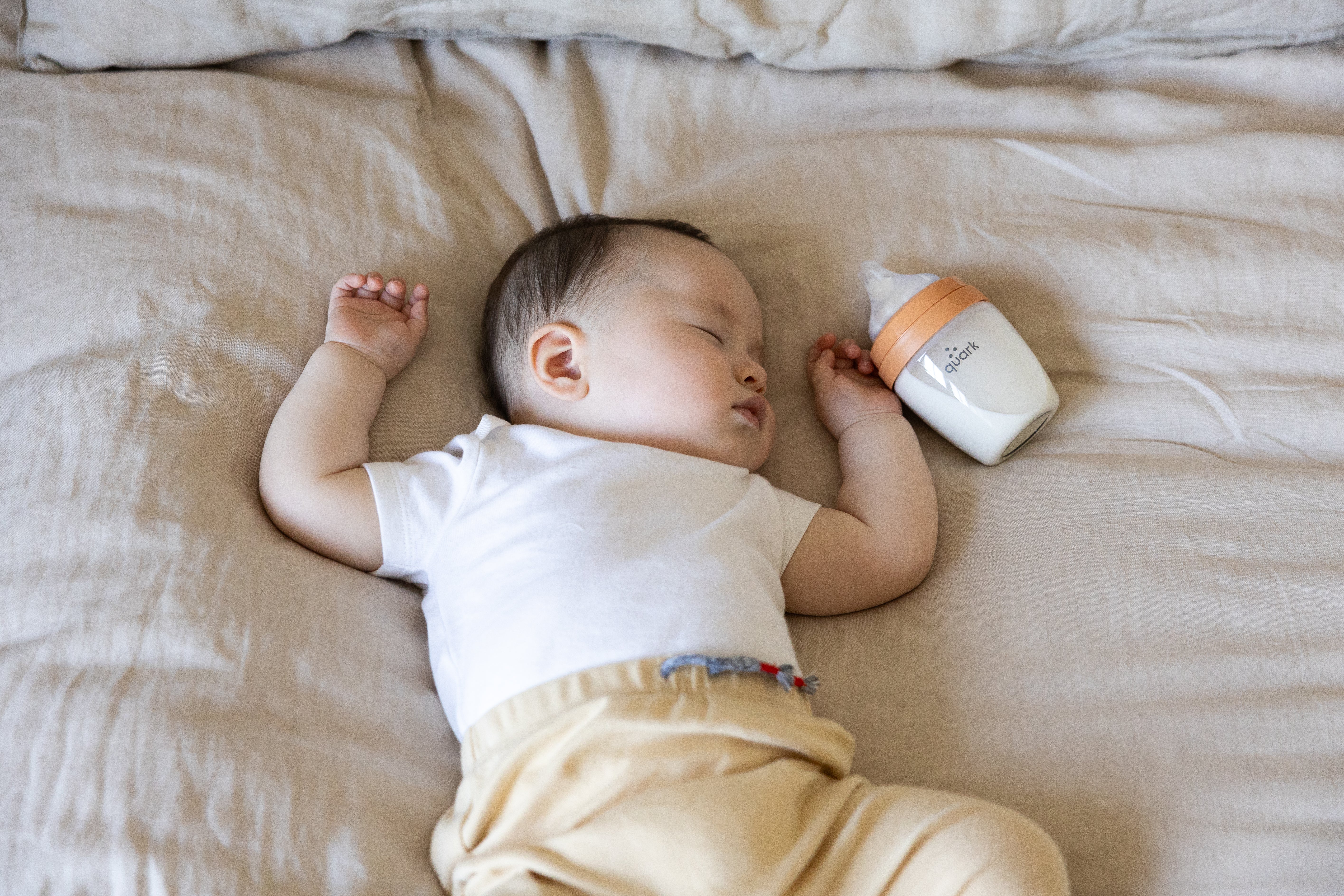 Sleeping newborn in white shirt next to Quark Baby bottle on beige bedding