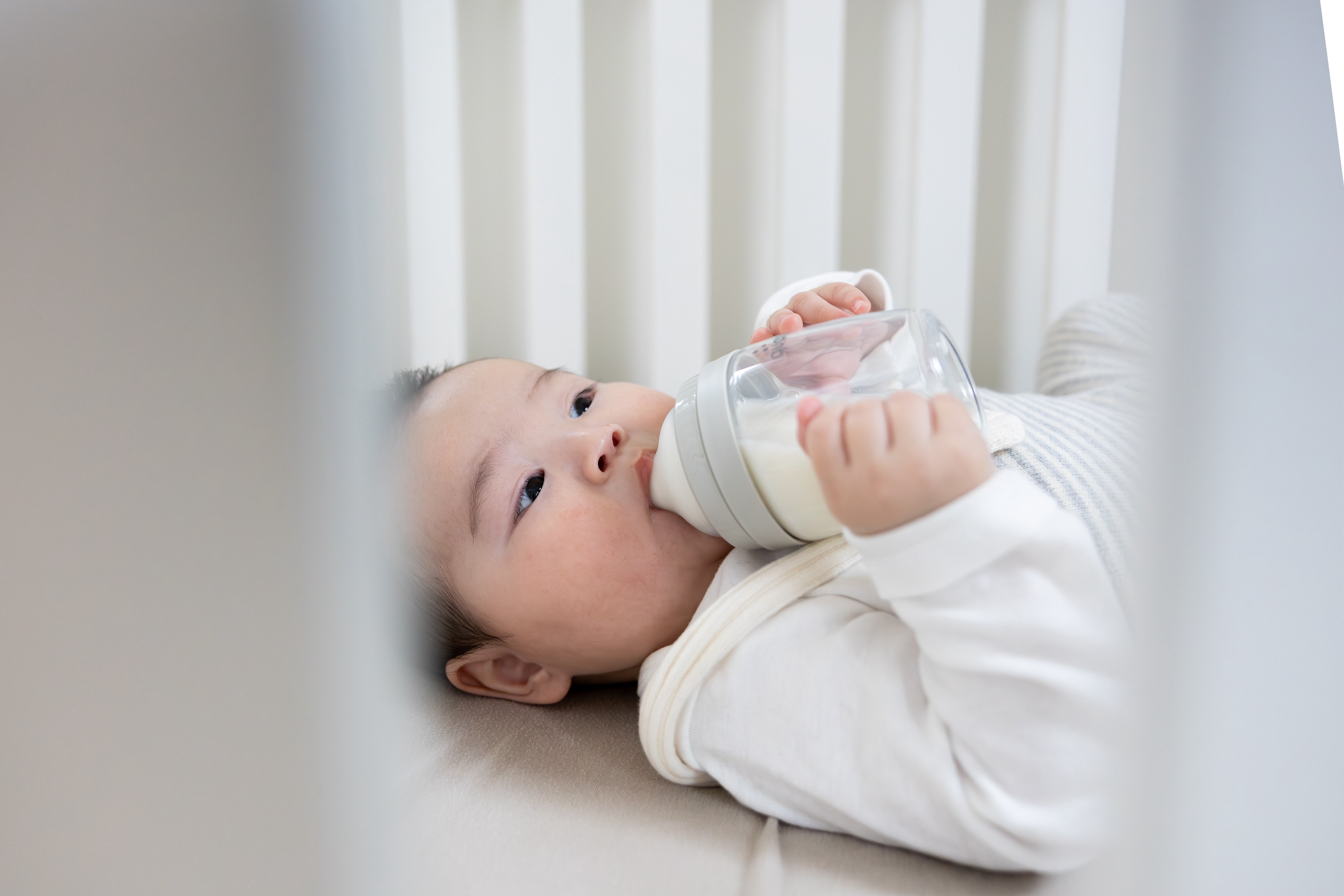 Newborn in white swaddle holding Quark Baby bottle with milk, lying in crib with vertical slats
