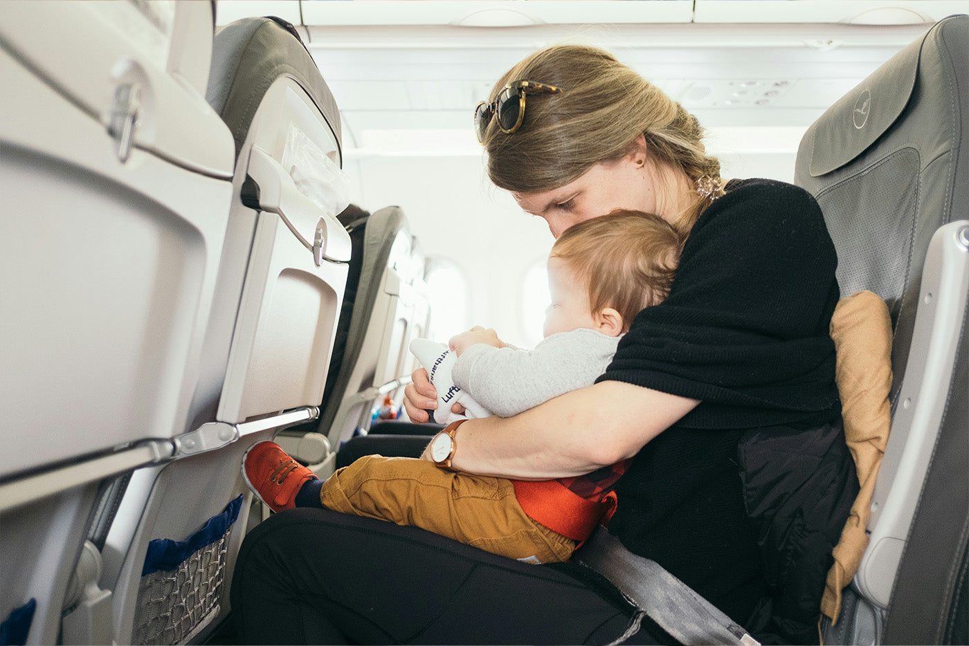 Mother holding toddler on airplane seat while infant drinks from Quark Baby bottle during flight
