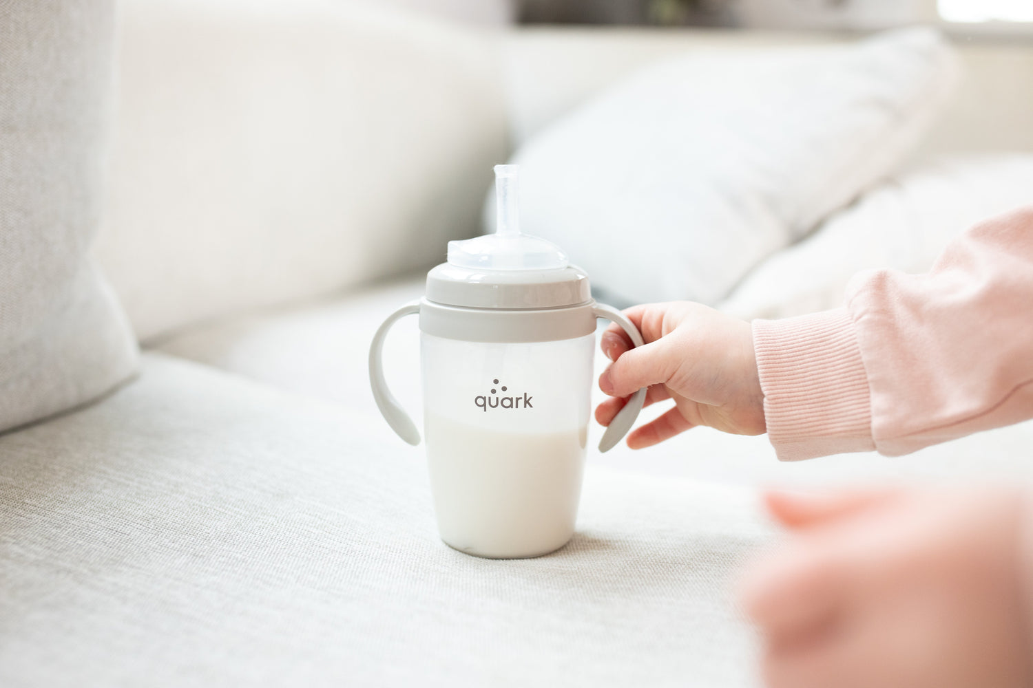 Adult hand presenting Quark Baby BuubiBottle sippy cup with handles on neutral nursery background.