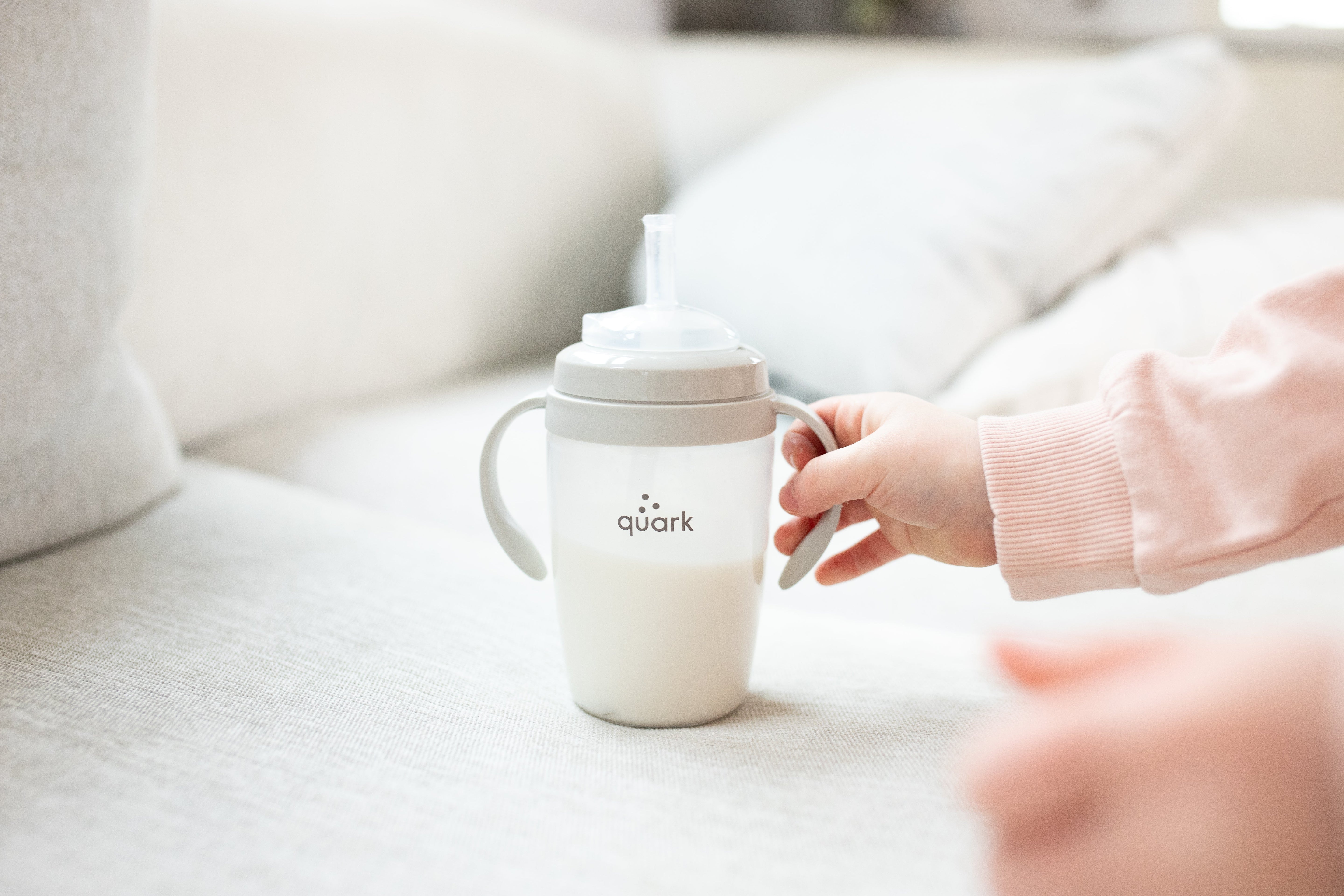 Adult hand presenting Quark Baby BuubiBottle sippy cup with handles on neutral nursery background.
