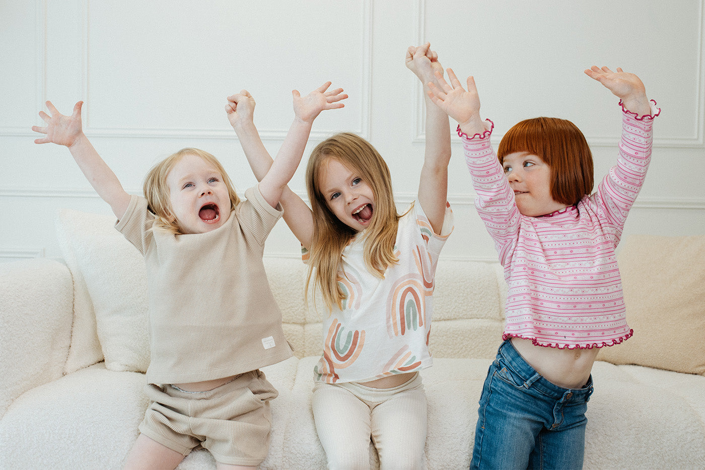 Three young children celebrating with raised arms on cream sofa, Quark Baby lifestyle shot.