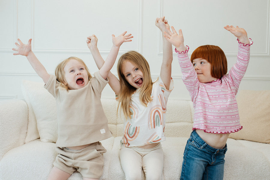 Three young children celebrating with raised arms on cream sofa, Quark Baby lifestyle shot.