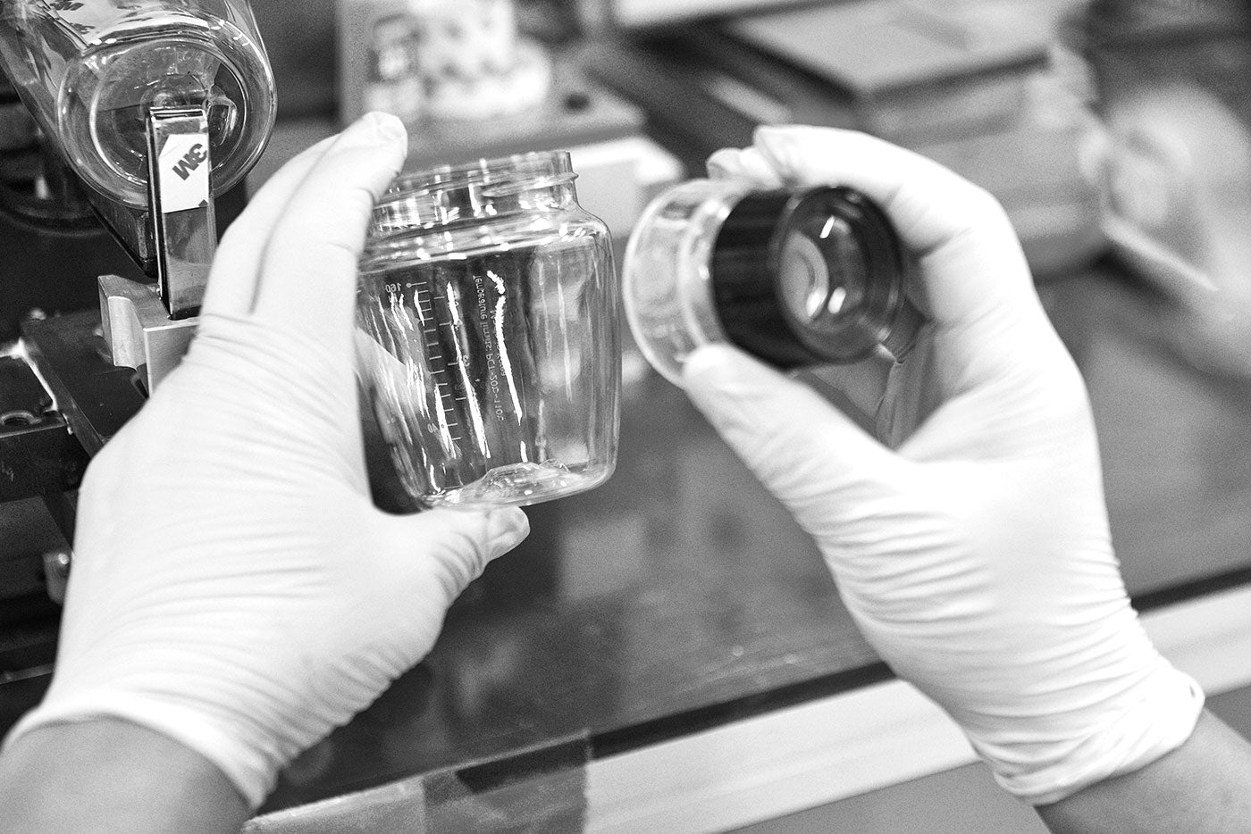 Hands in white gloves examining clear Quark Baby Tritan bottle and cap in laboratory setting.