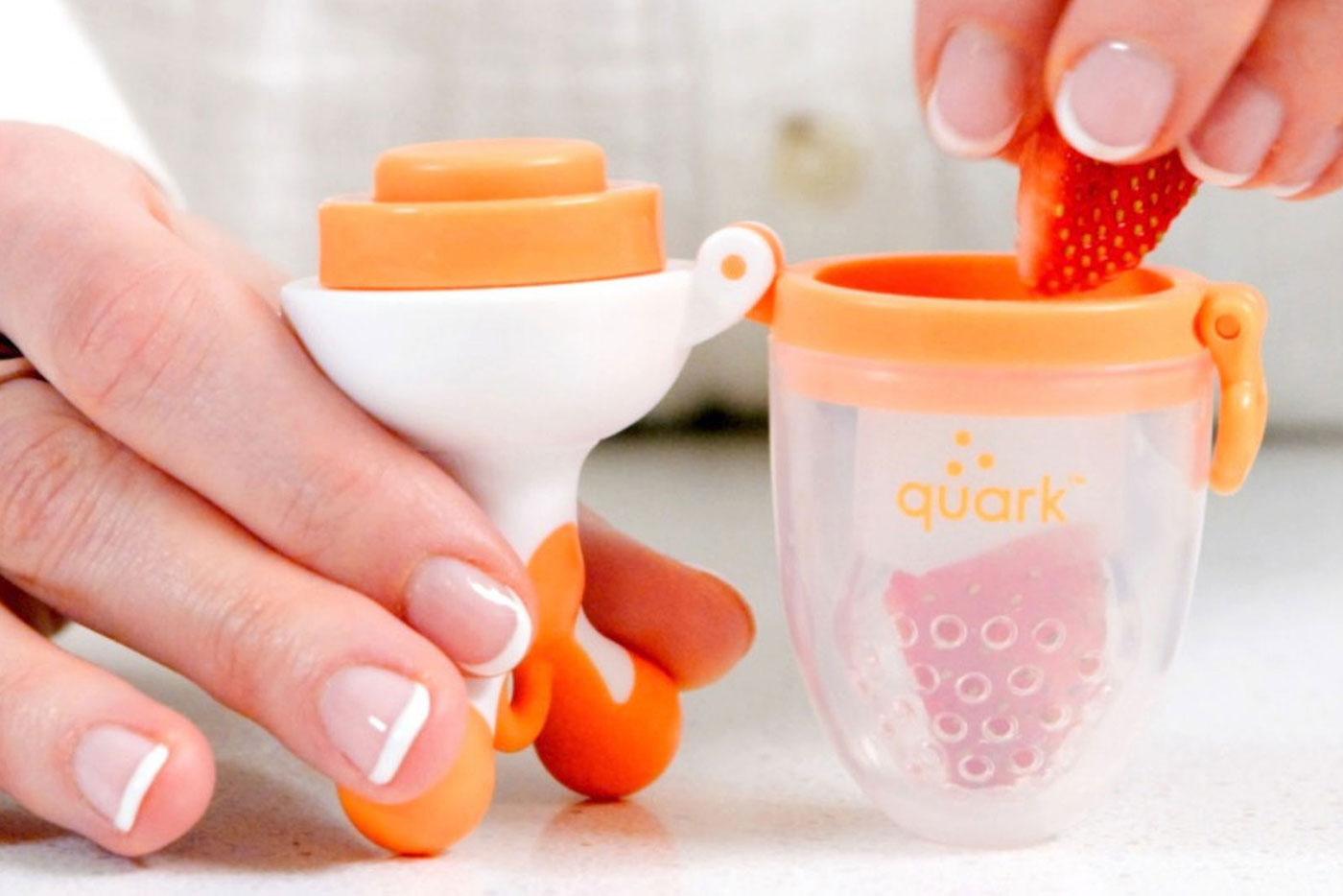 Quark Baby food feeder with strawberry being inserted into orange silicone pouch for first-year feeding.