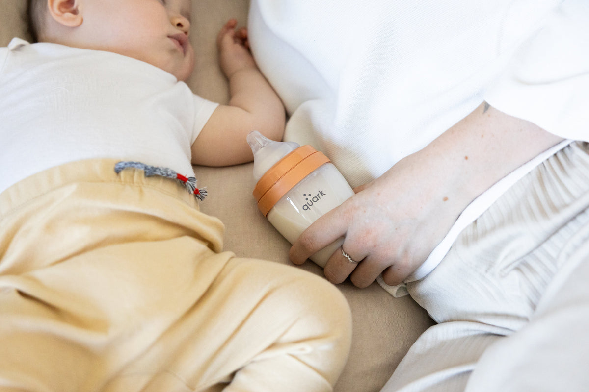 Adult holding orange BuubiBottle while paced feeding sleeping infant, demonstrating bottle feeding bonding technique.