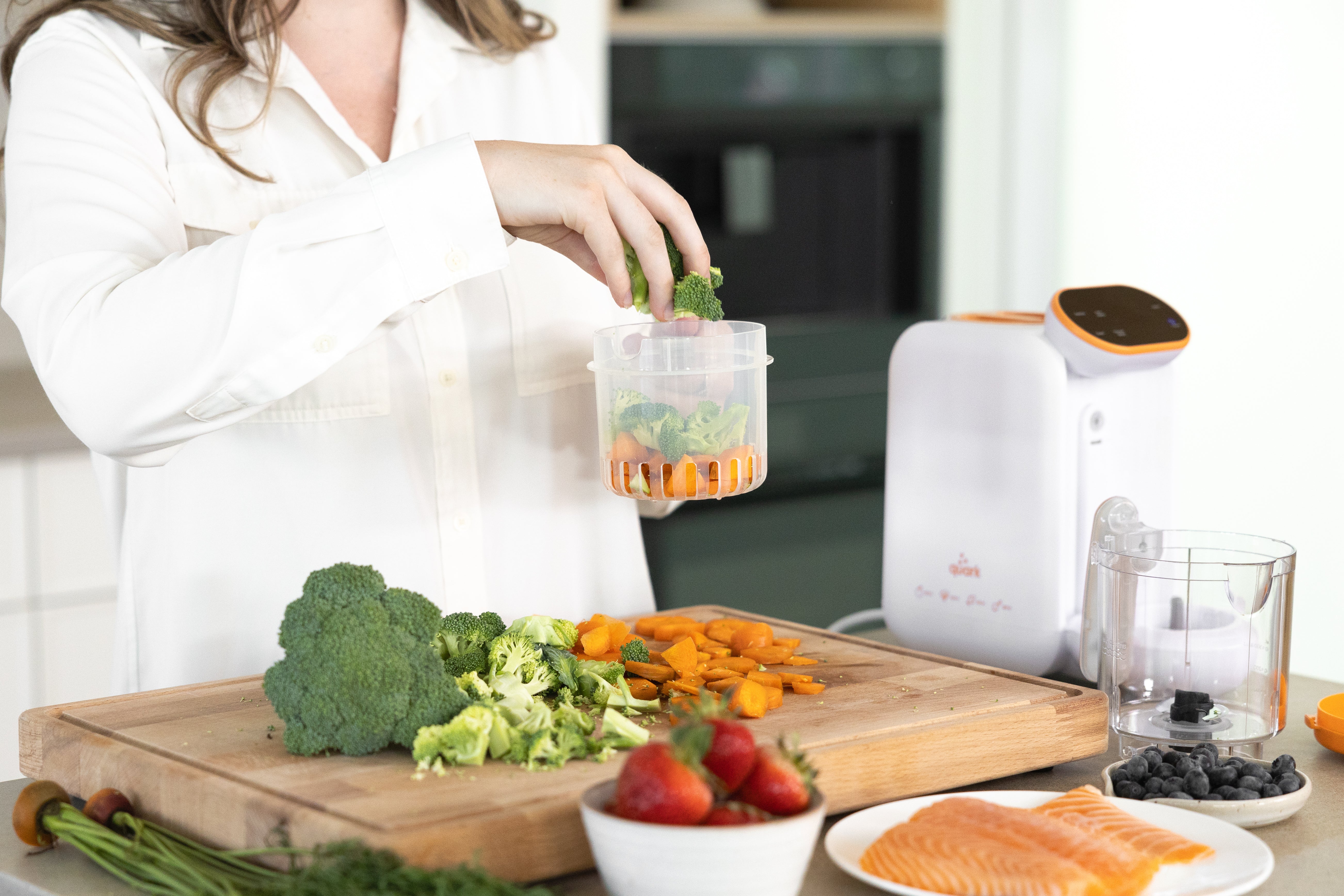 Woman adding broccoli to Quark Baby Quook blender with fresh vegetables and salmon on wooden board.