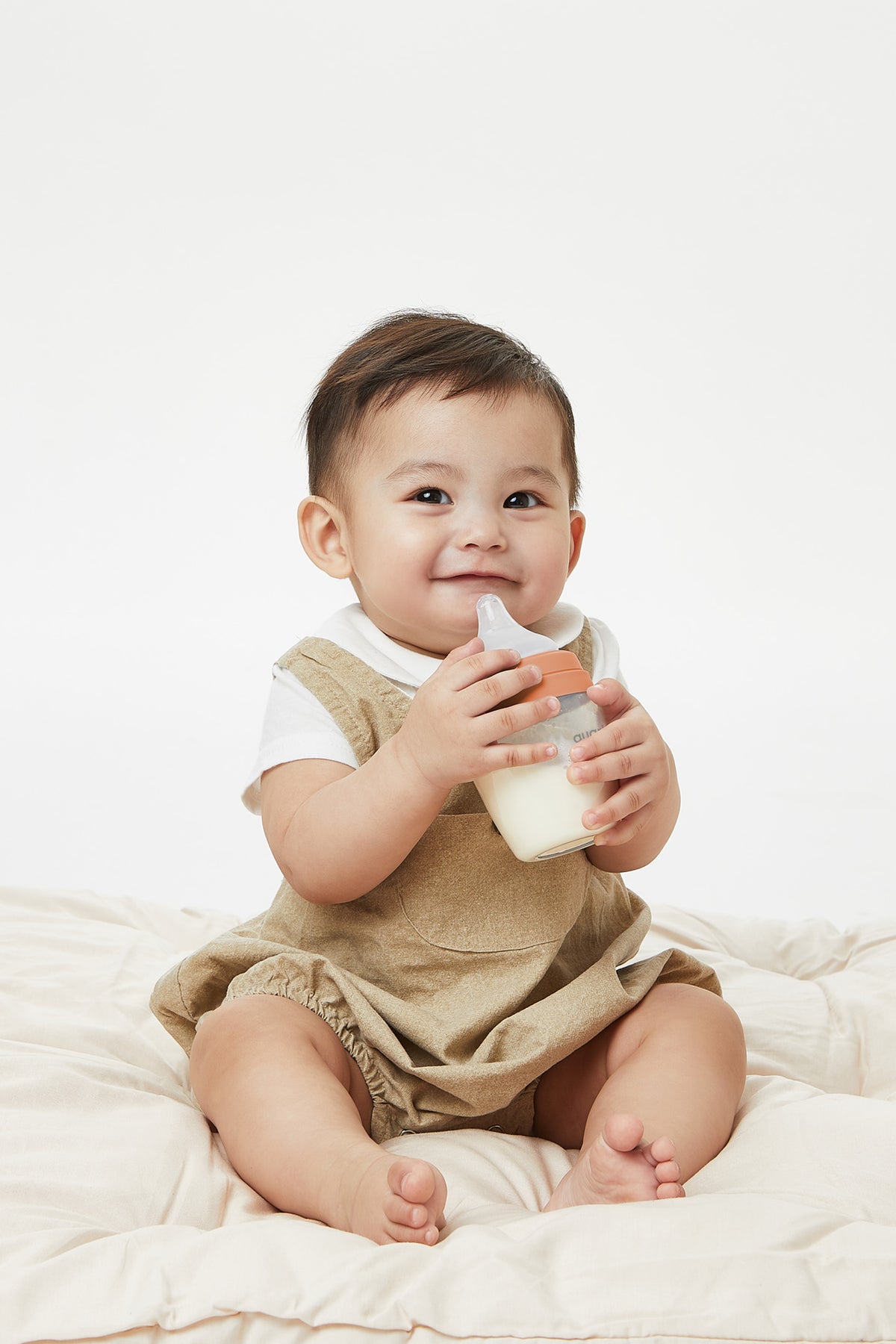Smiling baby holding Quark BuubiBottle Mini orange hybrid feeding bottle in beige outfit