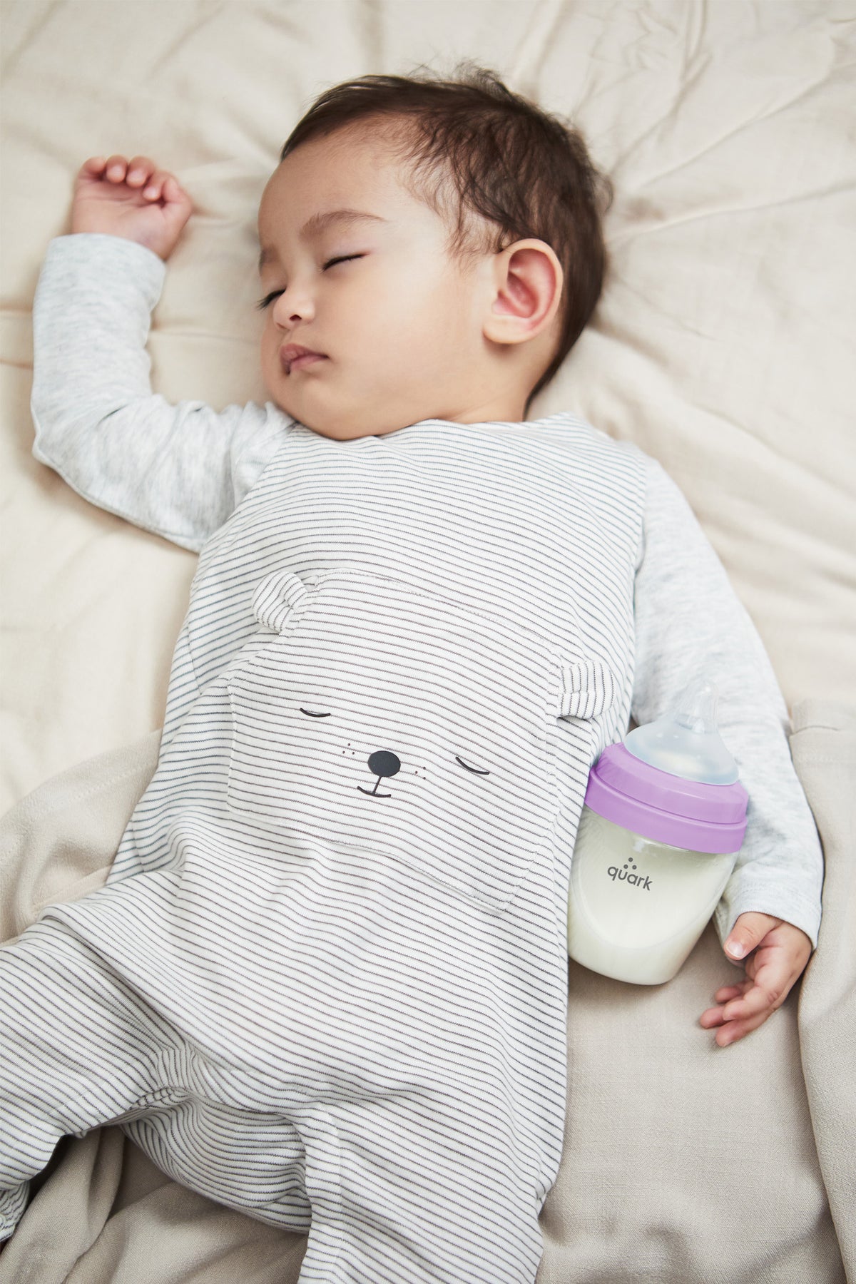 Sleeping baby in striped shirt holding Quark BuubiBottle Mini with purple cap on neutral bedding