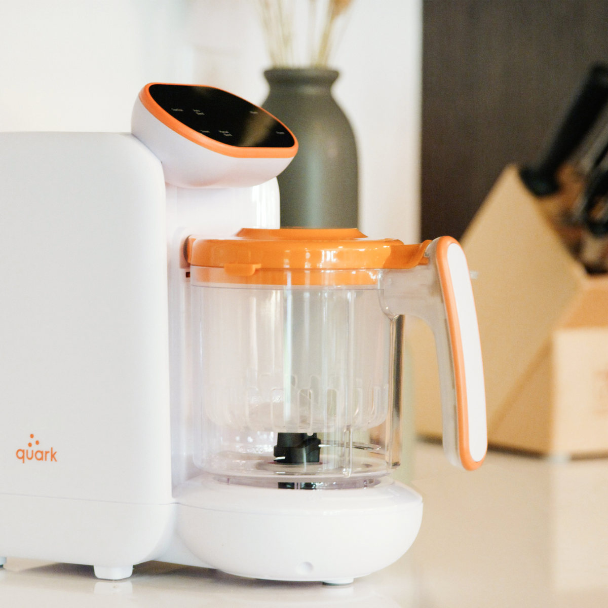 Quook baby food maker with orange accent lid on kitchen counter — compact 5-in-1 steamer blender for homemade baby food and bottle warming