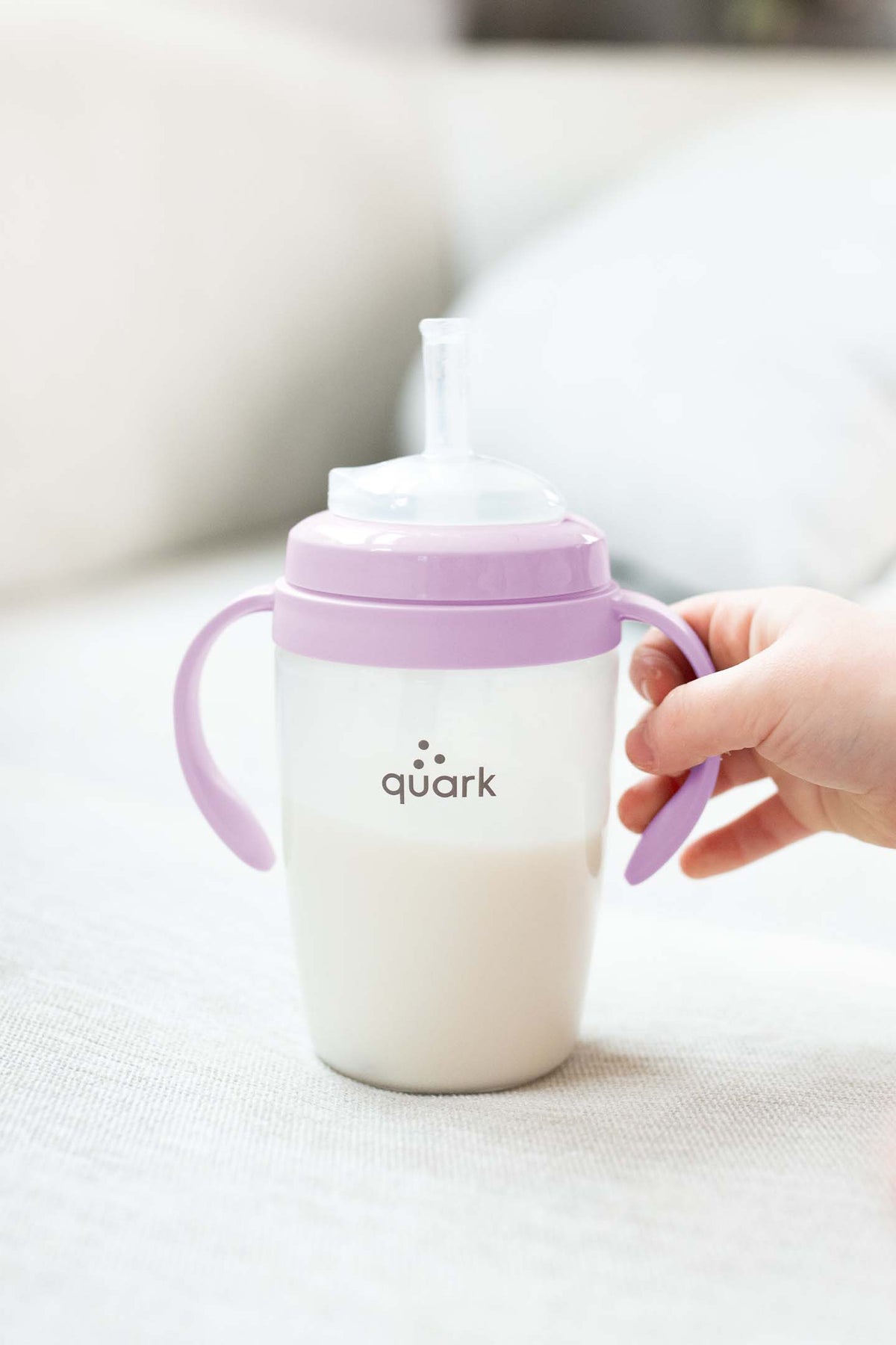 Toddler holding Quark BuubiBottle SipKit with purple handles and white spout on neutral background.