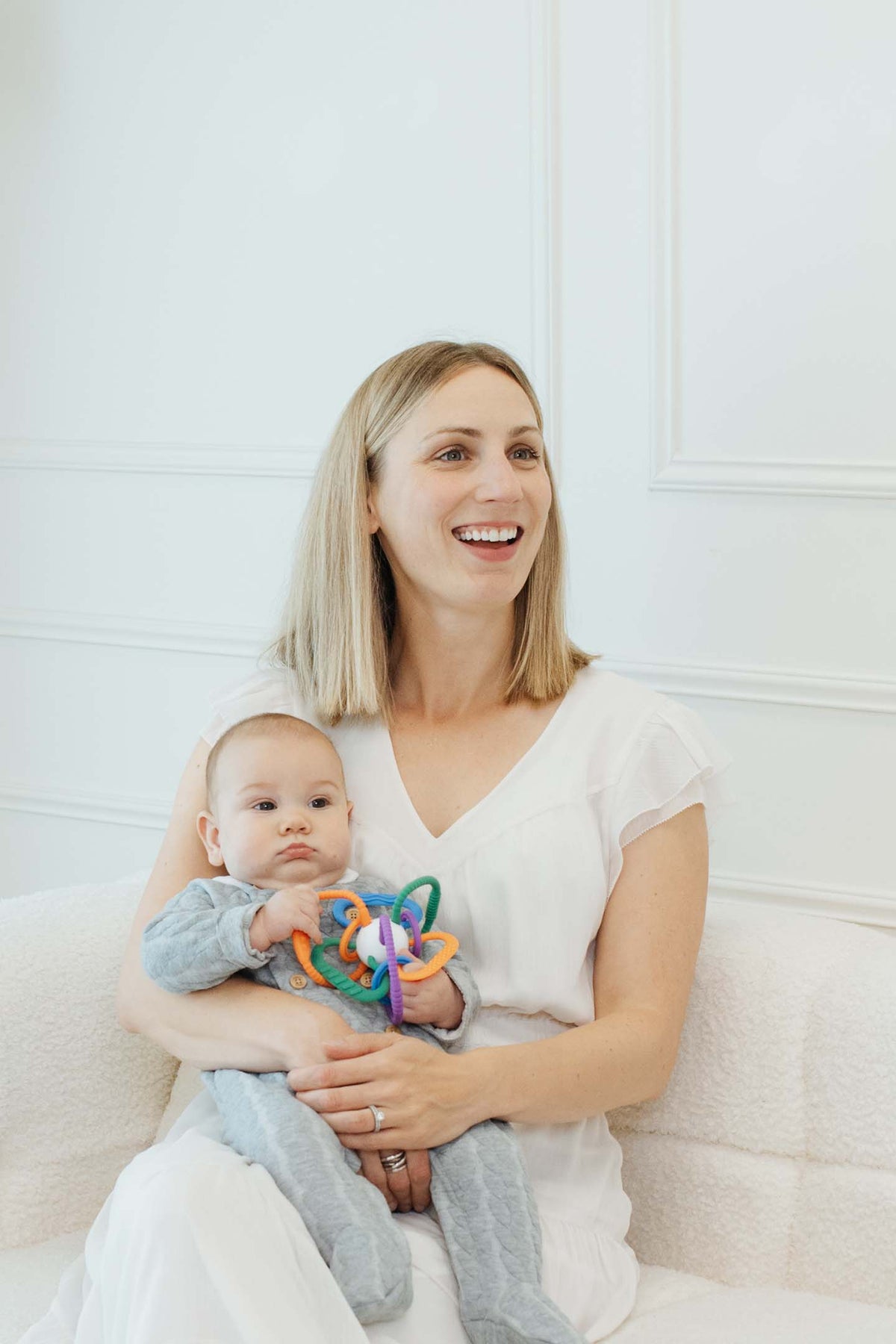 Smiling mother in white shirt holding infant with colorful Quark Thiingy sensory toy in lifestyle portrait