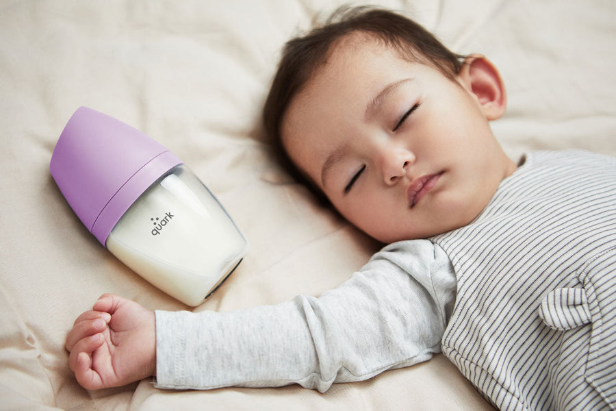 Sleeping infant beside Quark Baby purple bottle on cream bedding, feeding time lifestyle shot