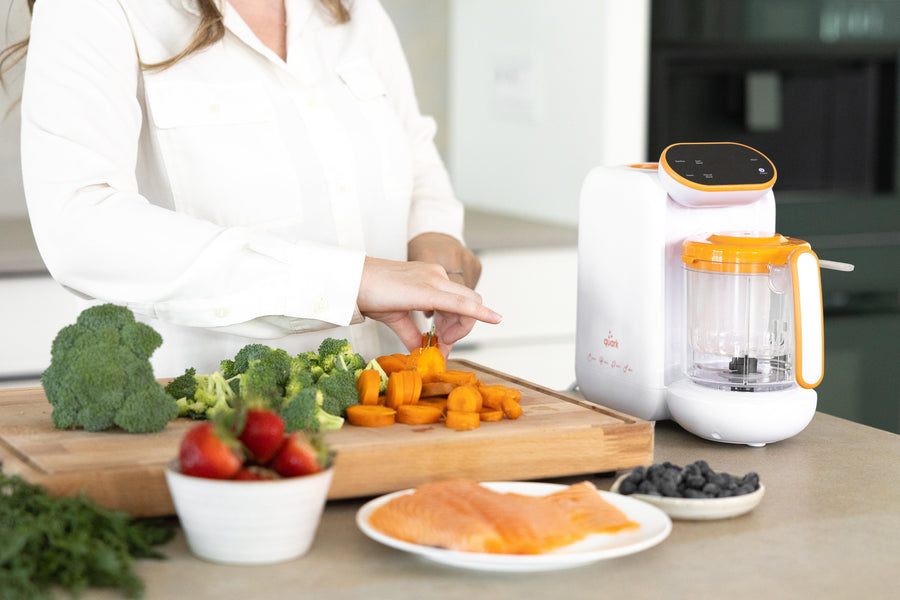 Woman preparing homemade baby food with Quark Baby Quook blender and fresh vegetables on cutting board.