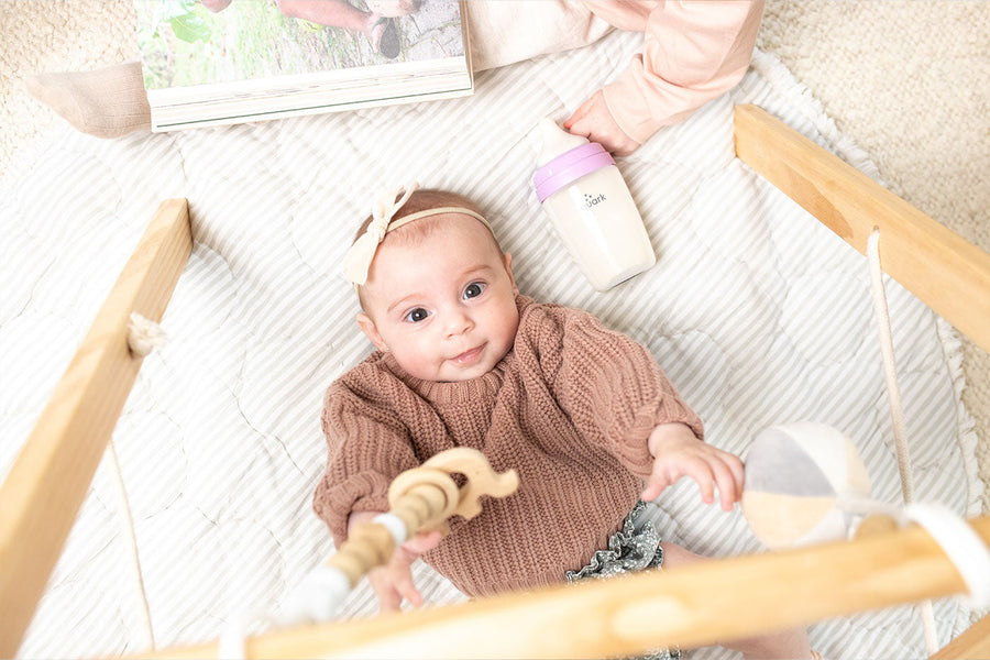 Baby in brown knit sweater with headband in crib with Quark Baby bottle and toys