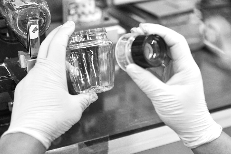 Hands in white gloves examining clear Quark Baby Tritan bottle and cap in laboratory setting.