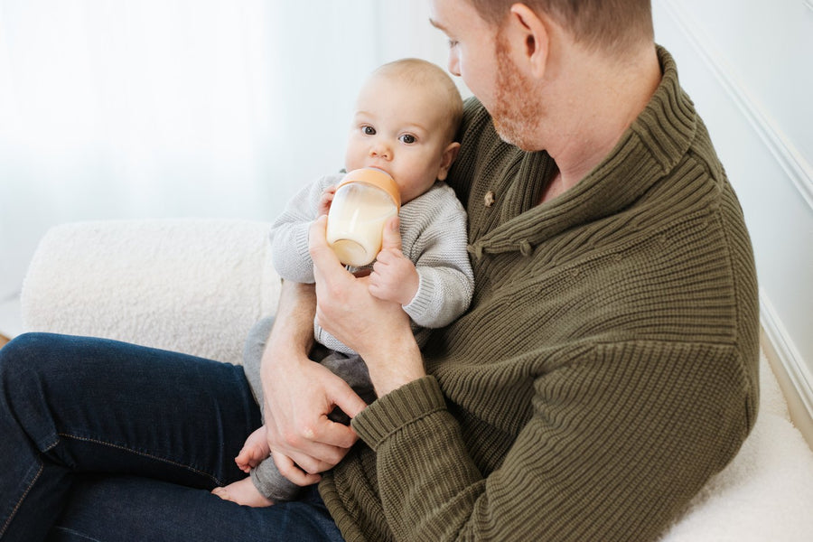 A father sitting on a sofa holding his baby while the baby drinks from a BuubiBottle