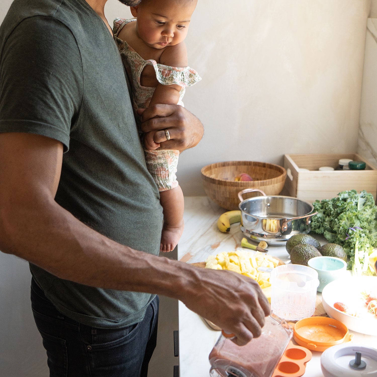 Adult holding infant while preparing colorful vegetables and fruit with Quark Baby mealtime tableware and storage.