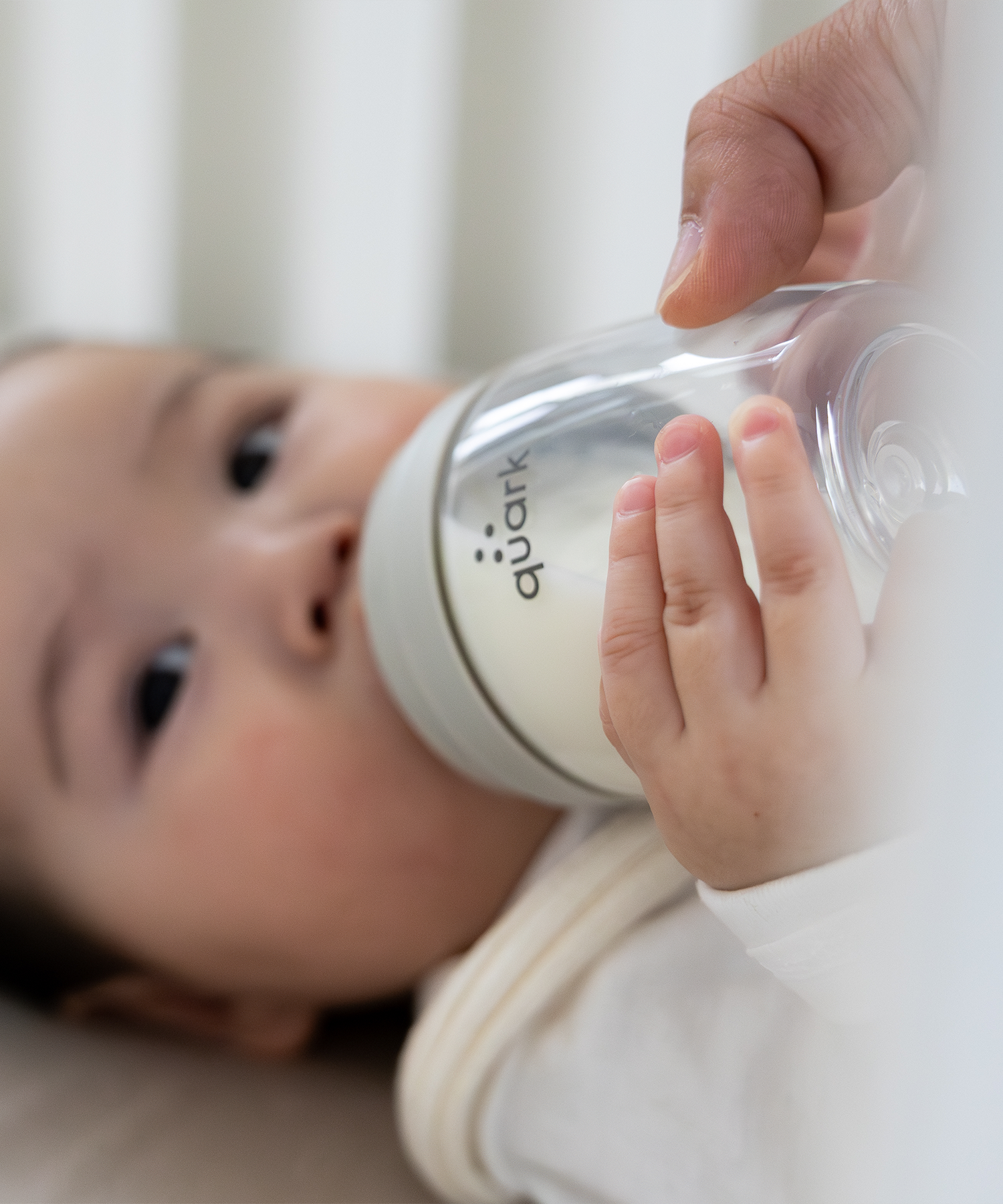Quark Baby bottle with white finish held by caregiver's hands while infant drinks, close-up view