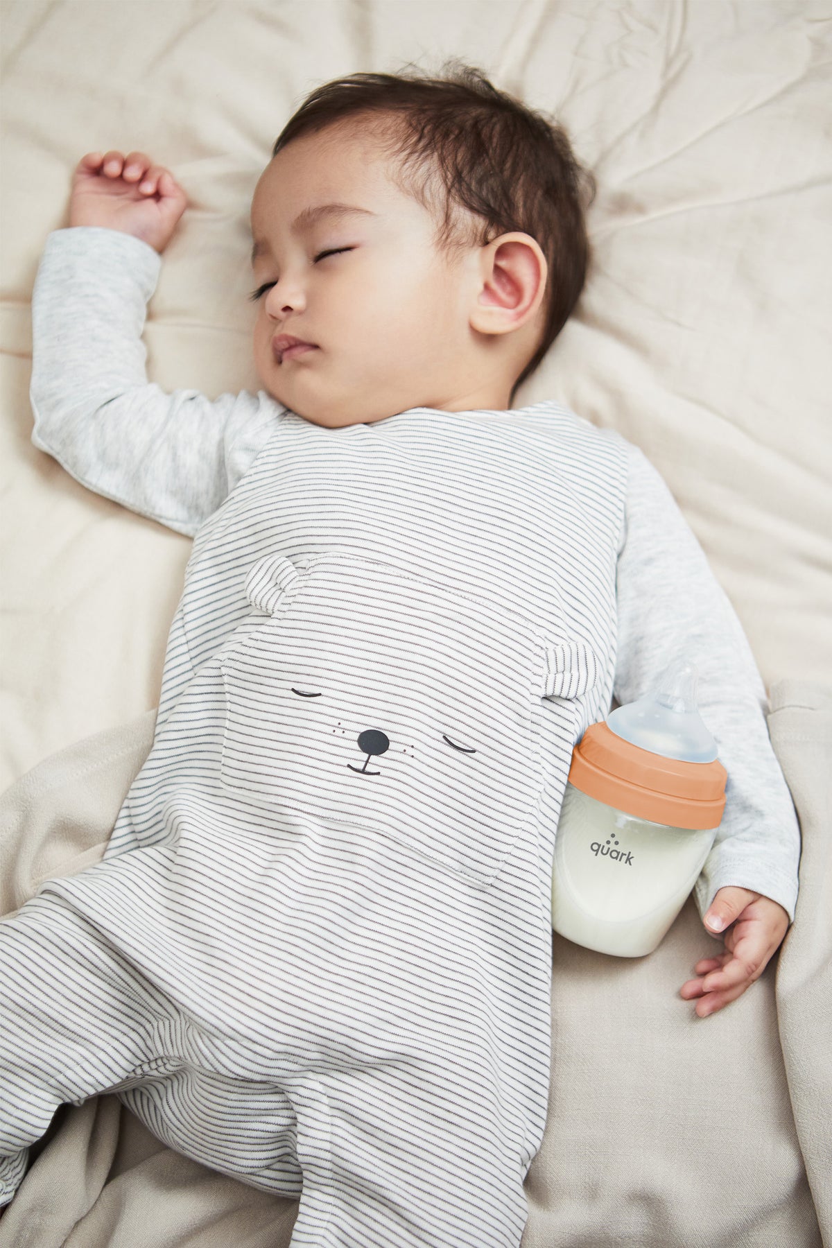 Sleeping toddler in striped shirt holding orange Quark BuubiBottle Mini with bear design on cream bedding