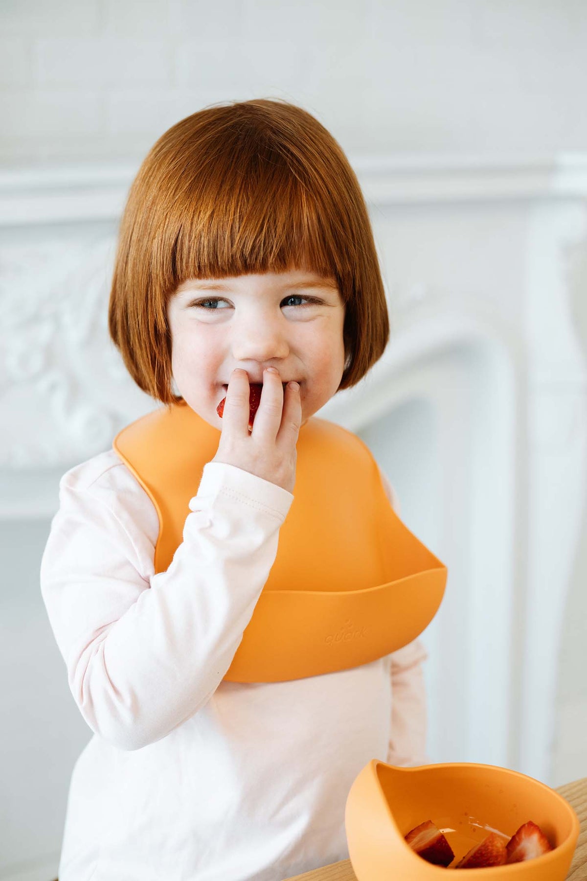 Red-haired toddler eating strawberries from Feedi orange silicone suction bowl with bib — toddler self-feeding, BPA-free silicone dining set