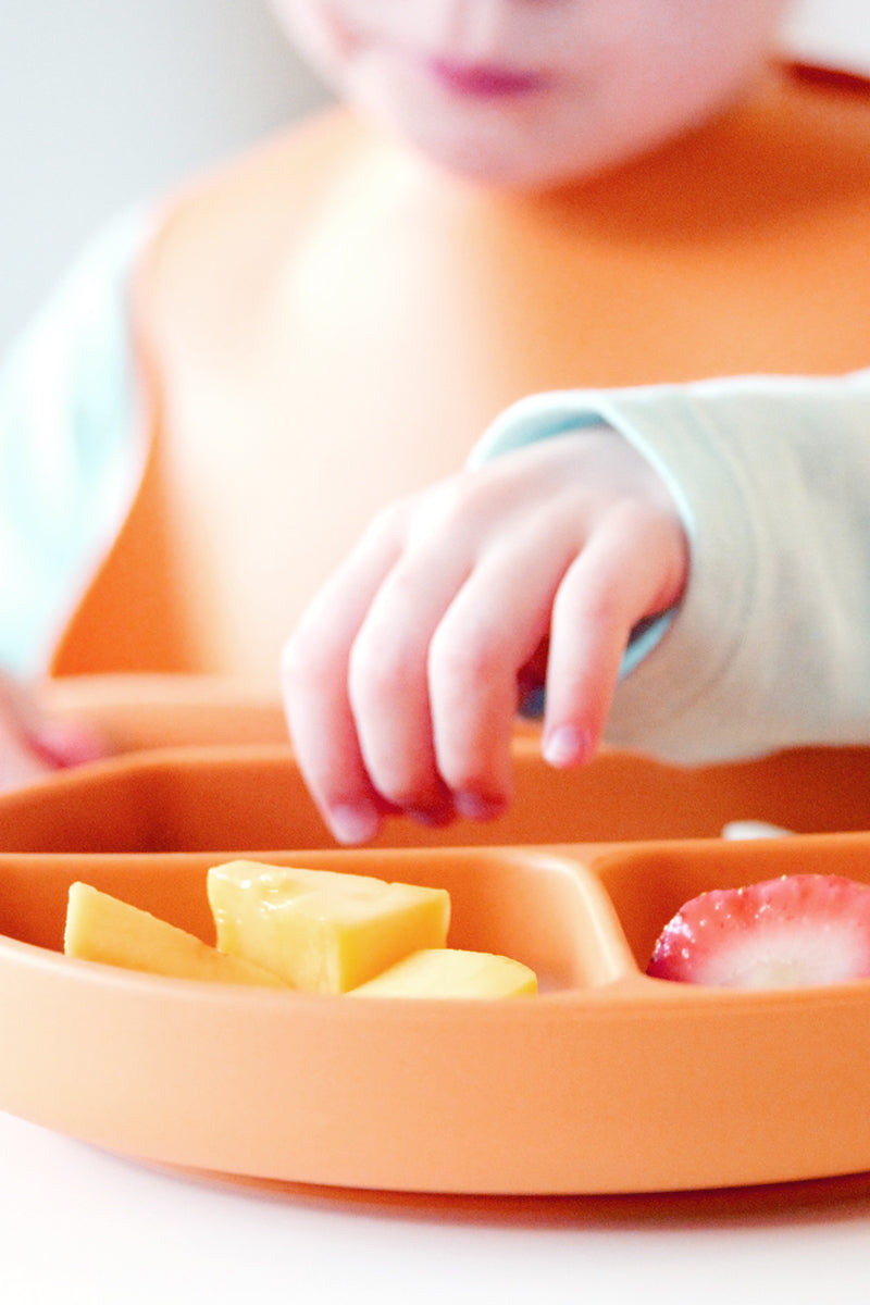 Toddler eating from Feedi orange silicone compartment plate and suction bowl — baby-led weaning dining set with soft spoon and bib