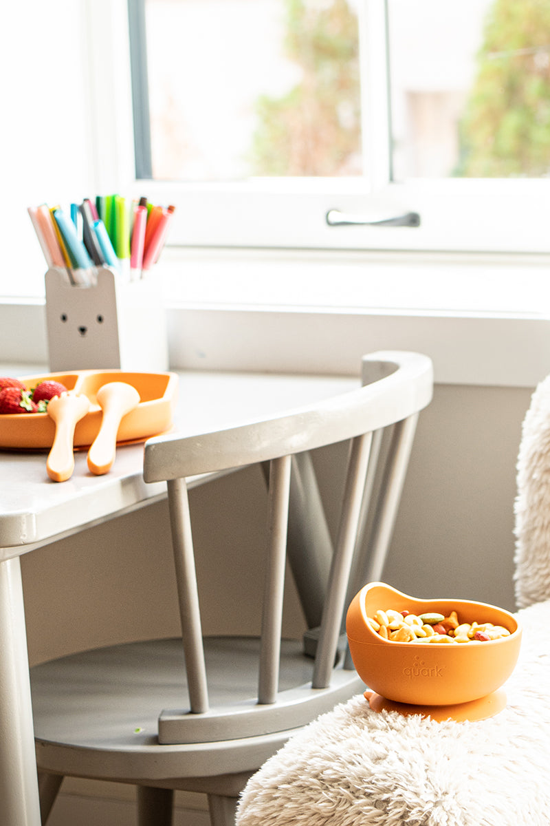 Feedi silicone baby dining set on white table near window — BPA-free orange compartment plate, suction bowl, and utensils for first solids
