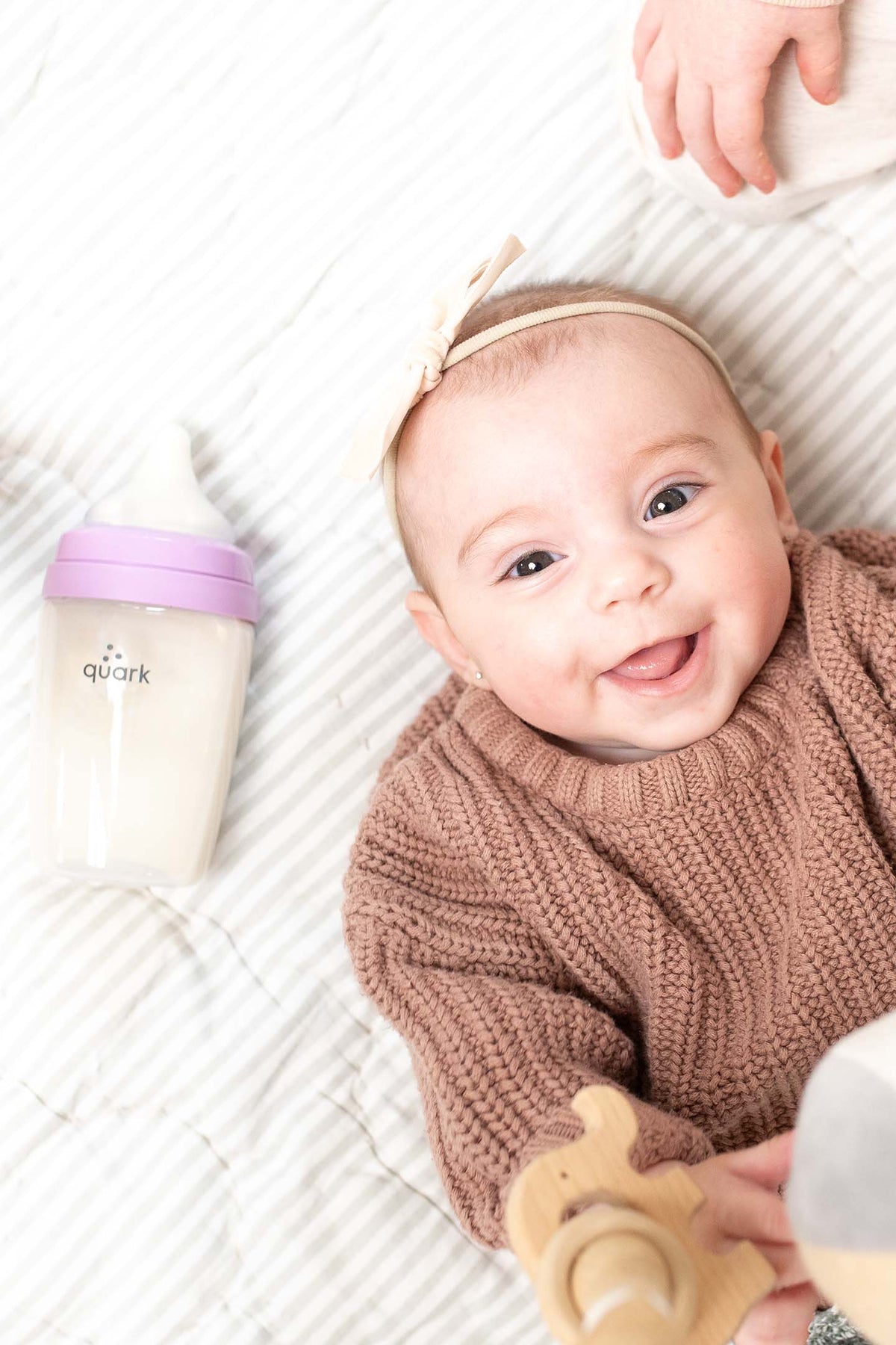 Smiling baby wearing brown knit sweater next to Quark BuubiBottle Max with purple cap on striped bedding