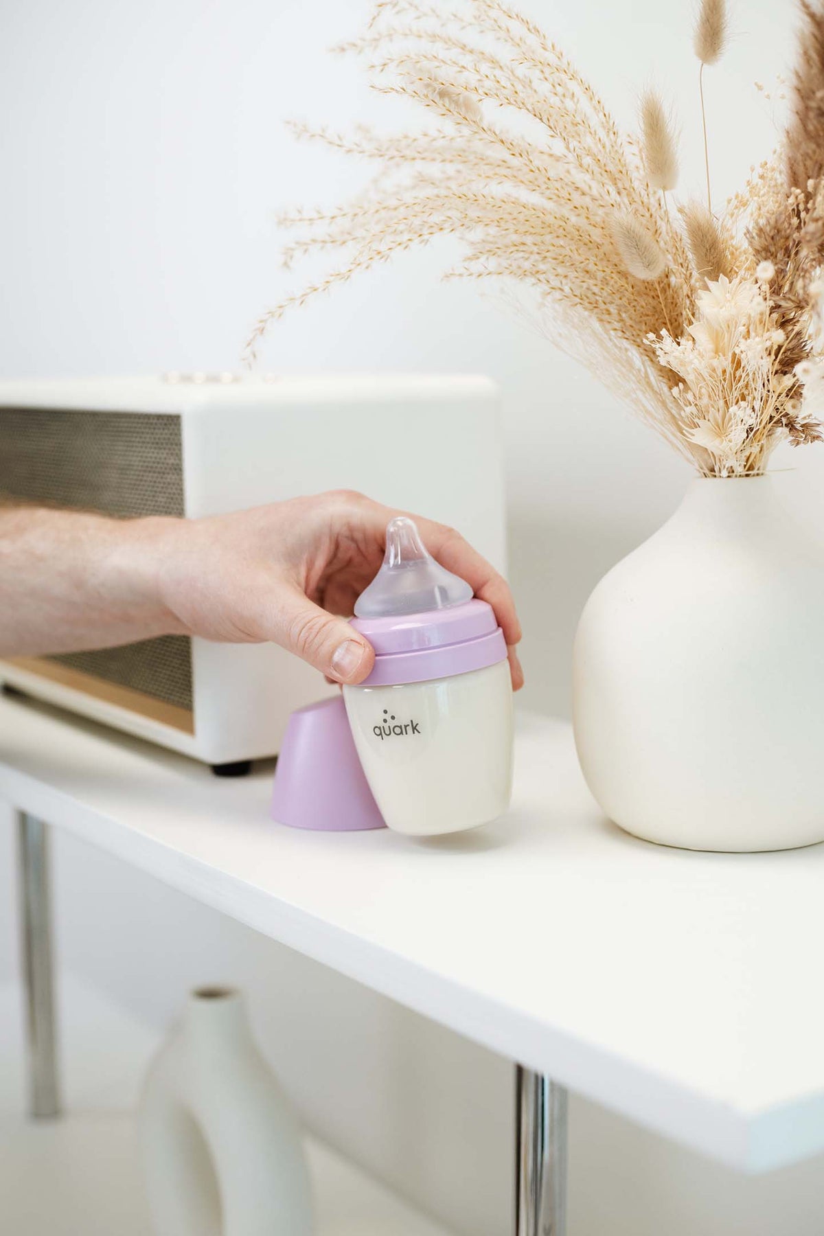 Adult hand holding Quark BuubiBottle Hybrid Feeding bottle on nursery shelf with dried florals