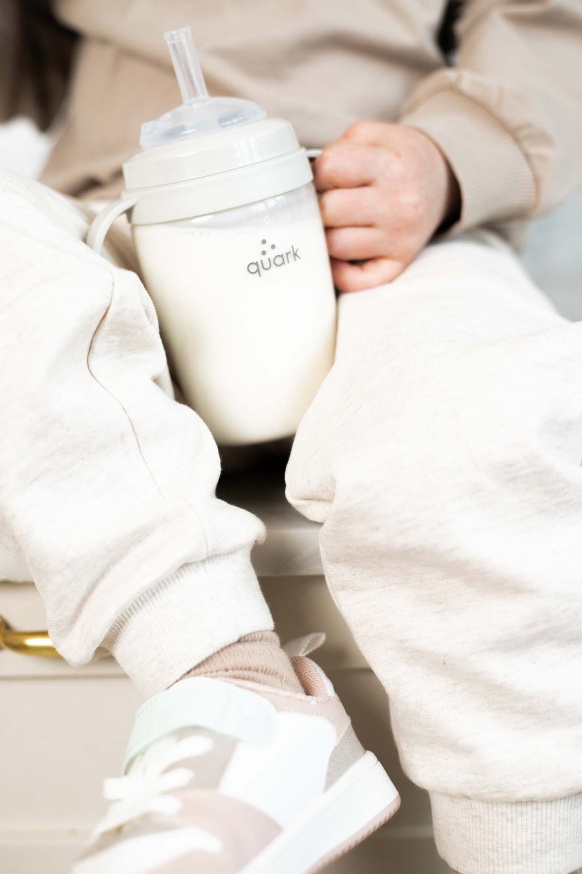Infant's hand grasping Quark BuubiBottle Sip cup with straw in cream knit clothing during daytime feeding moment