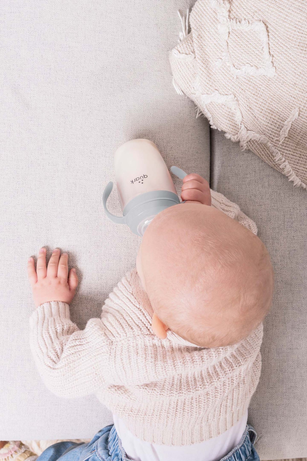 Baby in cream knit sweater holding Quark BuubiBottle SipKit conversion kit with grey band while seated on neutral fabric